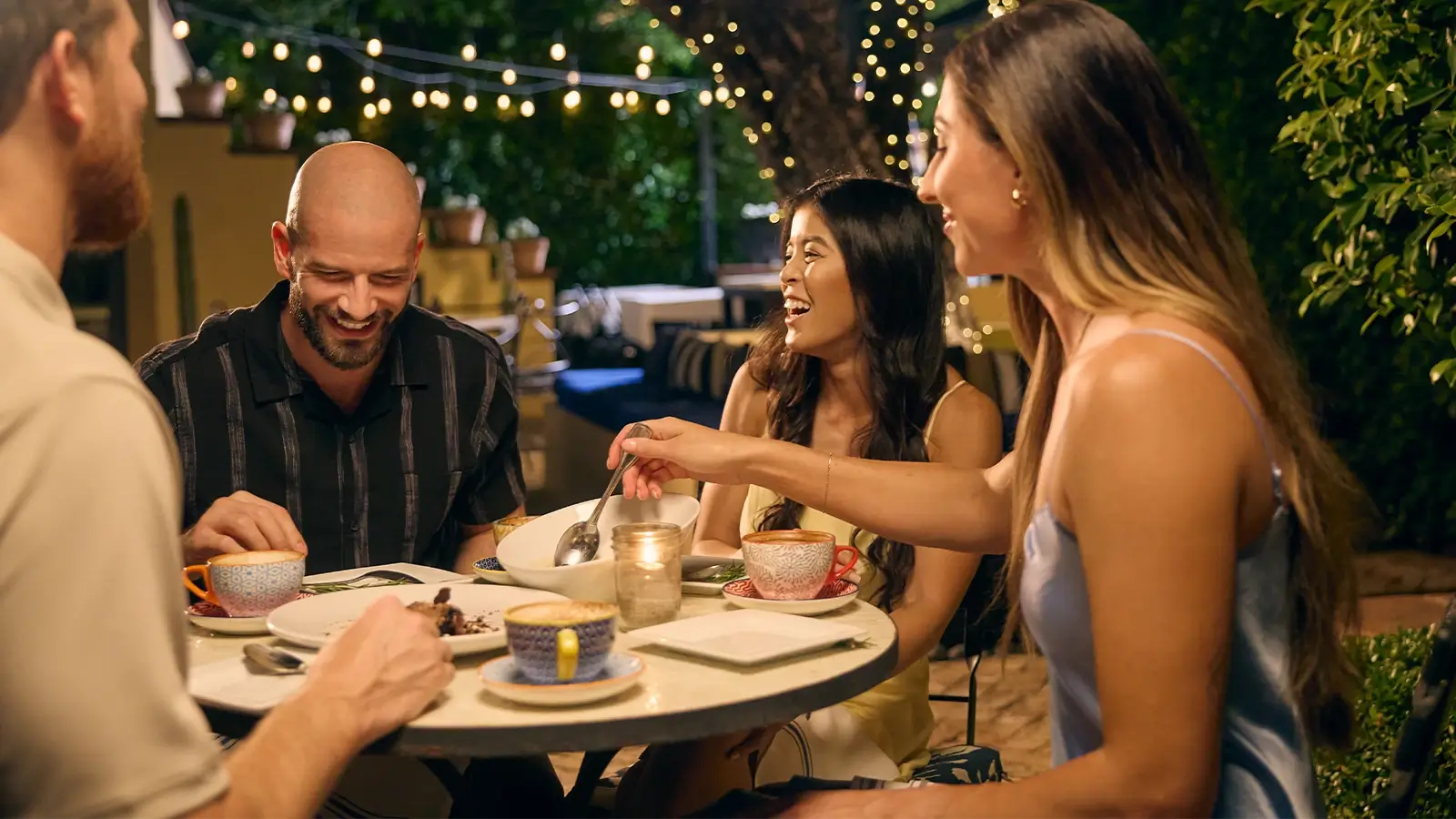 A group of four people are seated around a table, engaged in conversation at an outdoor restaurant. The warm lighting enhances the inviting atmosphere, with string lights adding a soft glow.